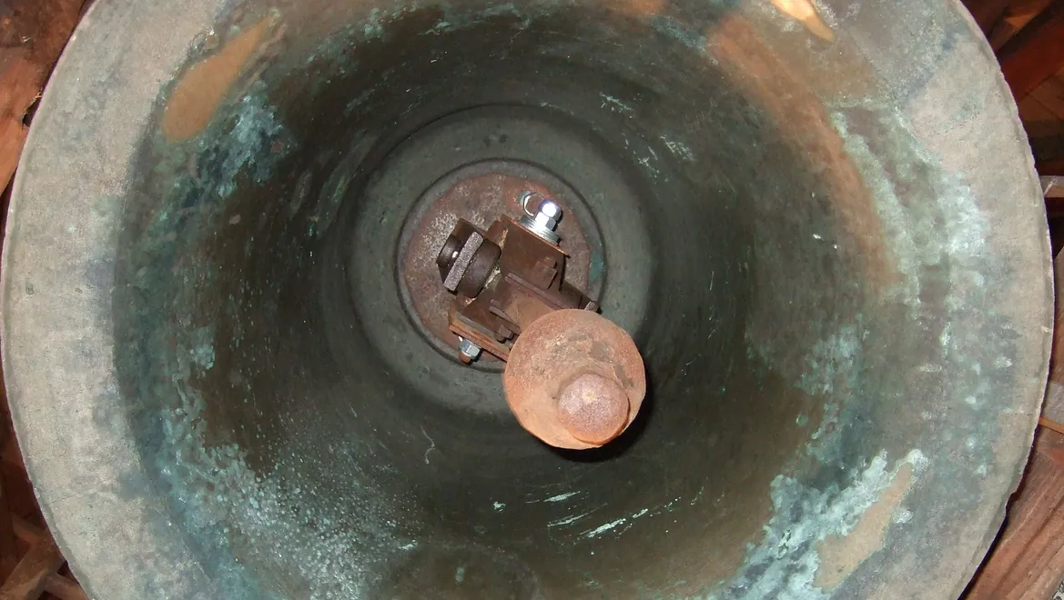 Das Innere der alten Kirchenglocke mit gut erkennbarer Klöppelmechanik. Es ist ein seltener Einblick in den technischen Aufbau einer Glocke, wie sie typischerweise in Kirchen hängt. Die Patina und Oxidationsspuren deuten darauf hin, dass die Glocke bereits seit vielen Jahrhunderten in Betrieb ist.