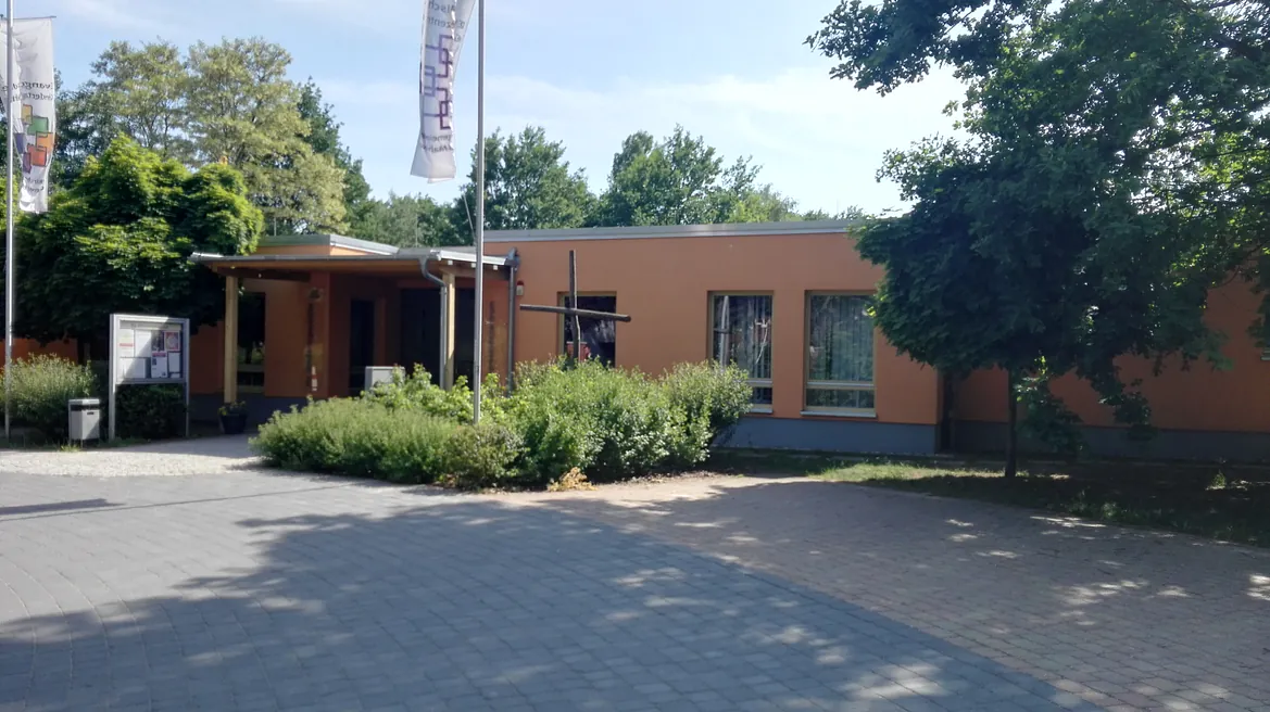 Auf dem Bild ist ein modernes, flach gebautes Gebäude mit orangefarbener Fassade zu sehen. ntrums Mahlow und der Kita vom Parkplatz aus gesehen.