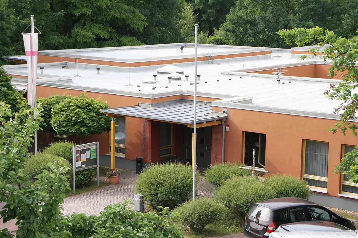 Das moderne evangelische Gemeindezentrum und ev. Kita, Bild vom Eingang mit Fahne, Kreuz, Schaukasten und Büschen und Bäumen.