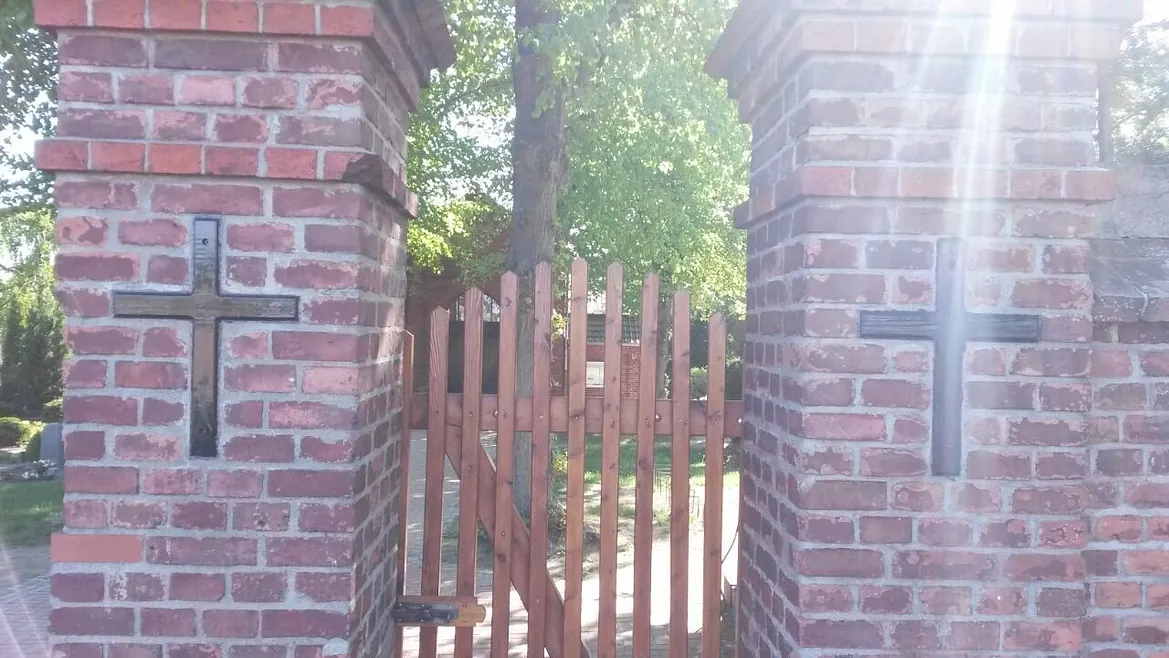 Holztor als Eingang zum Ev. Friedhof. Die Kreuze an den Pfeilern aus Backsteinsymbolisieren den christlichen Charakter des Ortes. Die ruhige, grüne Umgebung und das warme Licht verleihen dem Bild eine friedvolle und würdevolle Atmosphäre.